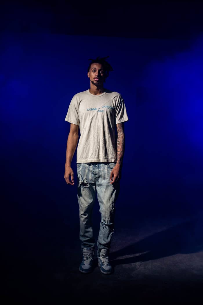 Kofi wearing a white T shirt and blue jeans against a blue and black background