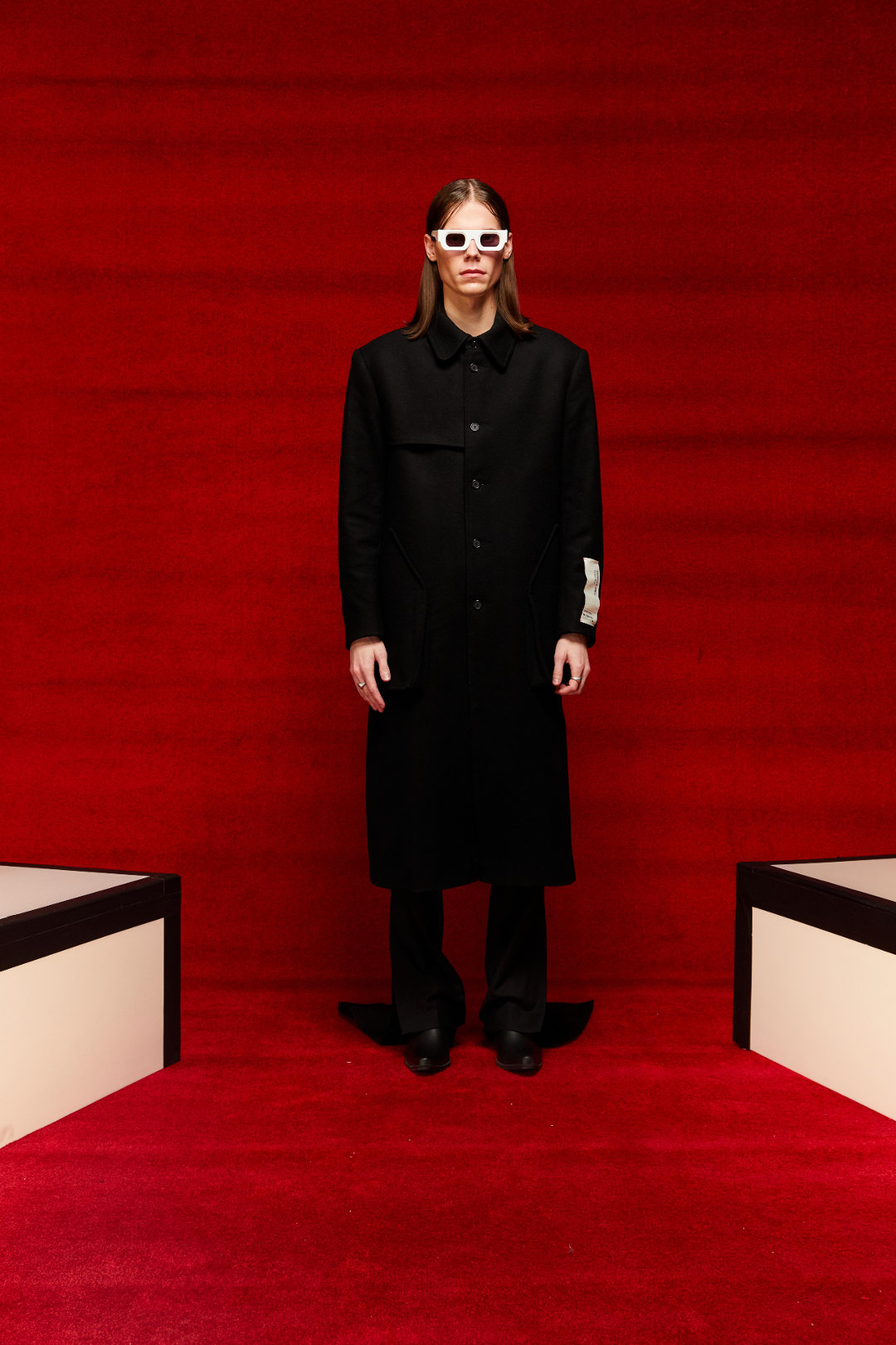 A person displaying a look from Mr. Saturday&#x27;s Fall/Winter 2022 collection. They are wearing a long black coat, black pants, and a black button up.