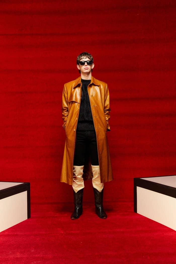 A person displaying a look from Mr. Saturday&#x27;s Fall/Winter 2022 collection.  They are wearing a long, warm brown leather jacket