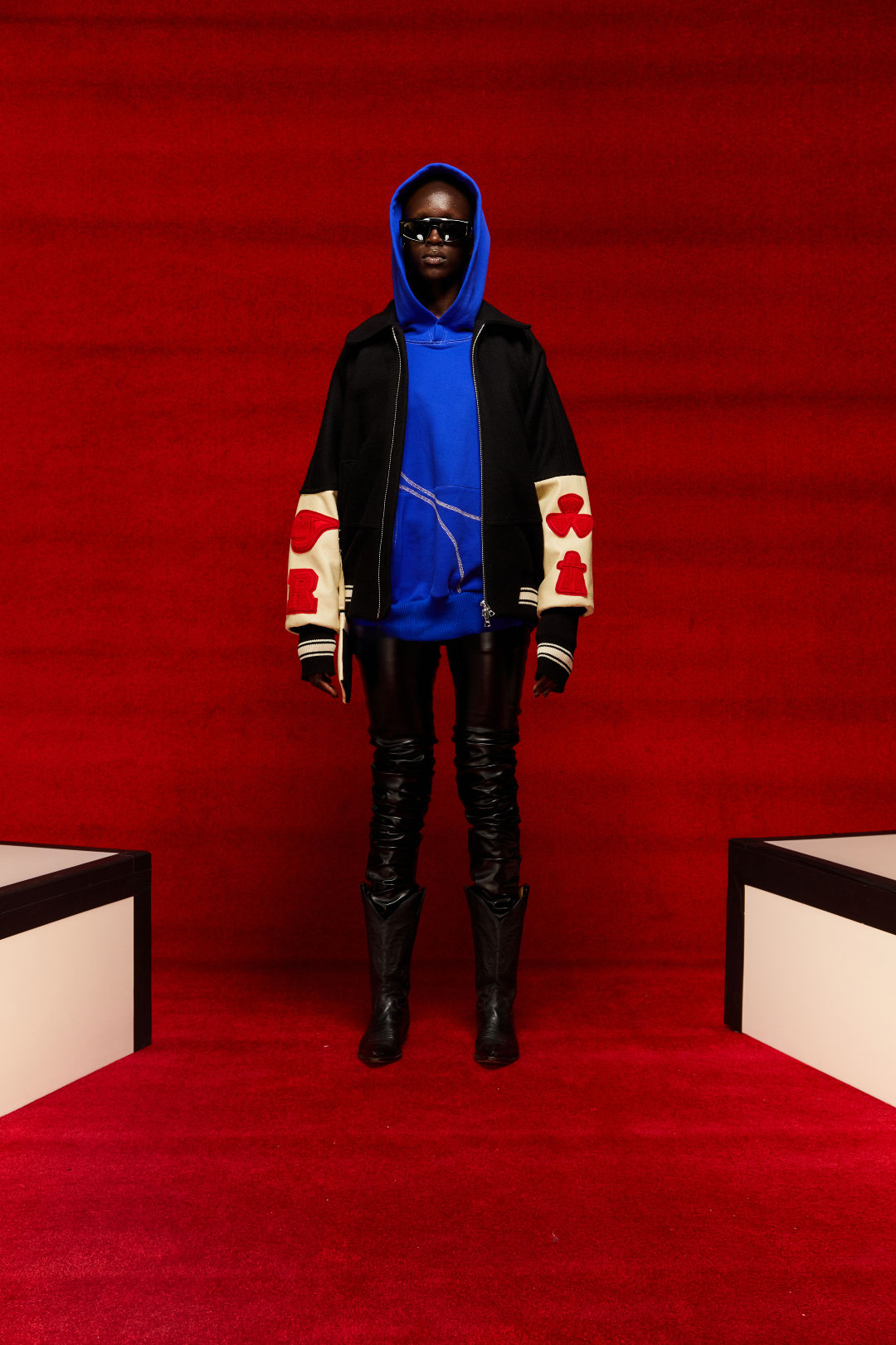 A person displaying a look from Mr. Saturday&#x27;s Fall/Winter 2022 collection. They are wearing a black varsity style jacket and a blue hoodie