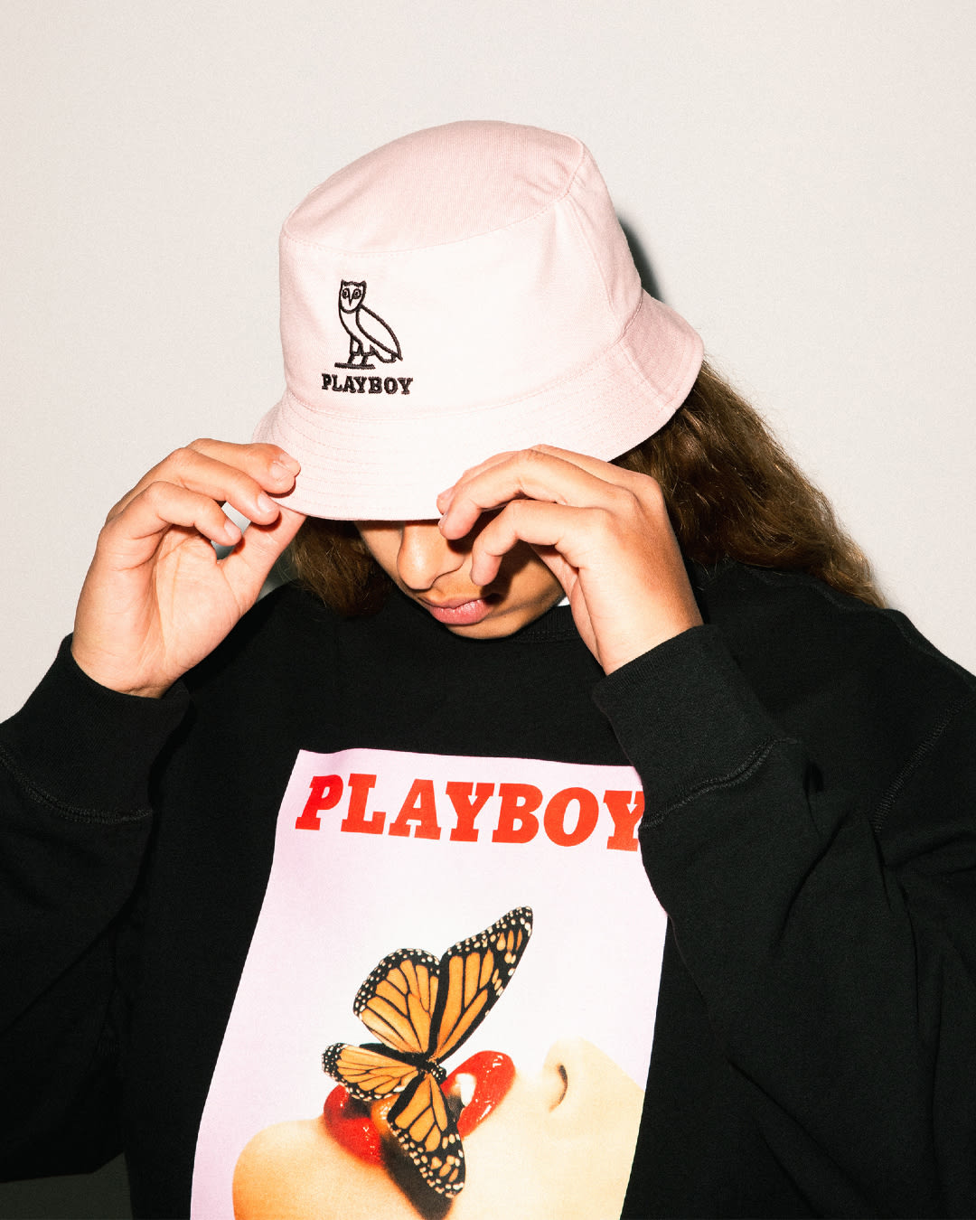 A model wearing a pink bucket hat with Playboy and OVO branding. He is also wearing a black crewneck