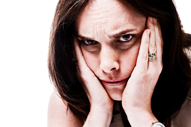 Woman of fair skintone with dark circles under her eyes, looks angry and confused as she holds her face in her hands