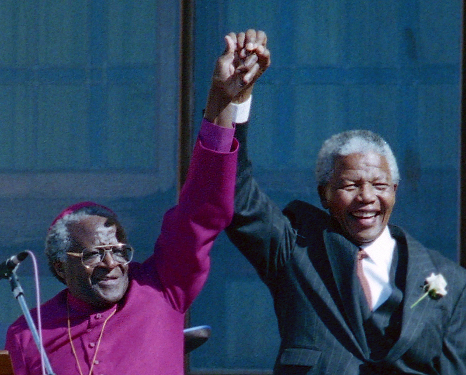 A smiling Tutu and Mandela stand together with raised hands clasped 