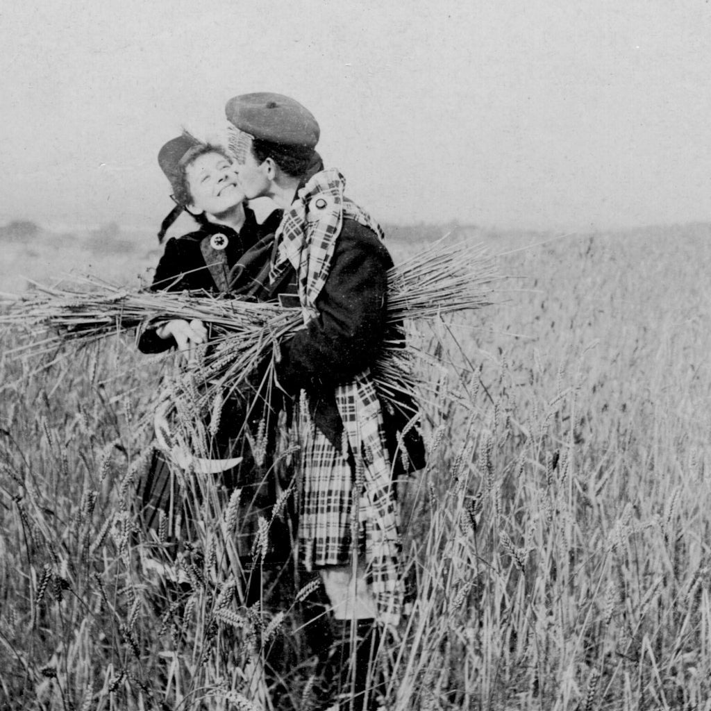 Un hombre vistiendo un kilt besa a una chica en un campo de centeno. Alrededor de 1910.
