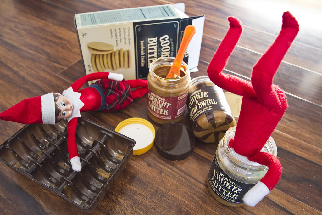 Jars of cookie butter combos and cookies, with one elf lying down on top of a packet of cookies and the other with their entire head in a jar