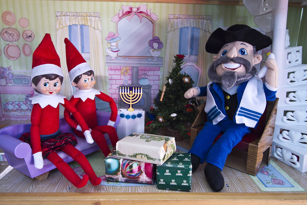 Two elves sitting on a couch opposite a man wearing a Jewish prayer shawl and a menorah between them
