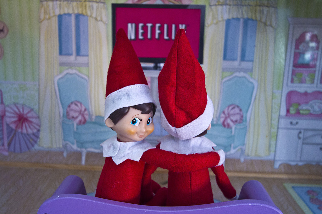 Two elves sitting in a seat, with one putting their arm around the other and turning around and looking at the camera