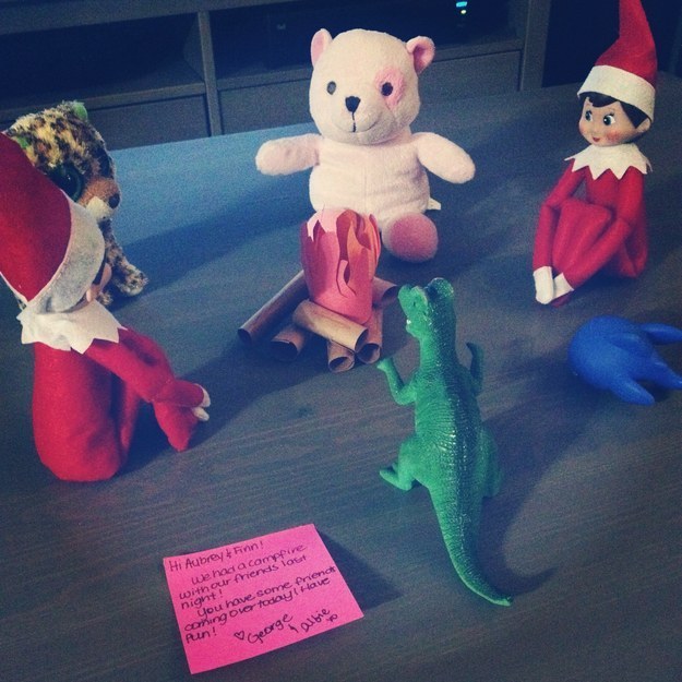 Two elves sitting in a circle with a dinosaur and a teddy bear, with a paper &quot;campfire&quot; in the middle, and a Post-it note referring to a campfire they had with their friends last night