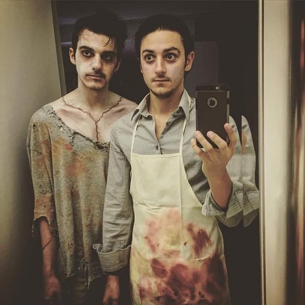 One man with a pale face and ripped clothing and one man wearing a bloody apron