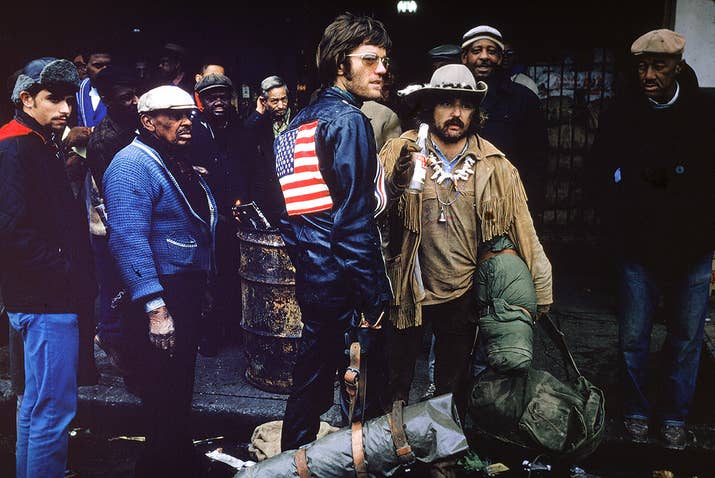 Dennis Hopper holds a bottle in his hand as he directs a setup for the film Easy Rider in New Orleans. Actor Peter Fonda stands next to him in sunglasses and motorcycle leathers while a group of extras and onlookers mill about behind them.