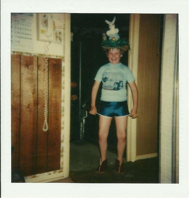 A smiling boy in short satin shorts and a large bonnet standing in a doorway