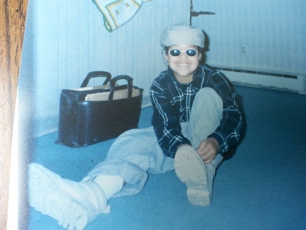 A smiling child wearing loose jeans, a flannel shirt, a cap, and sunglasses and sitting on a rug