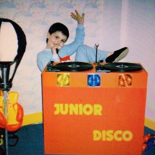 Child sitting at a &quot;Junior Disco&quot; booth with records on top and holding a microphone