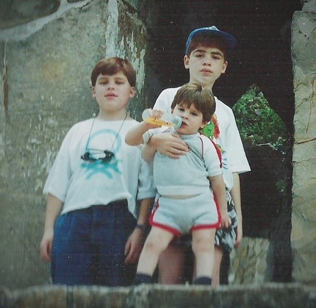 A toddler in shorts and a bottle in their mouth posing in front of two older youths