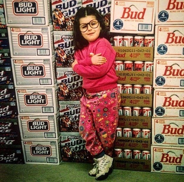 A child with glasses and shoulder-length hair posing in front of stacks of Bud Light and other beers in a store display