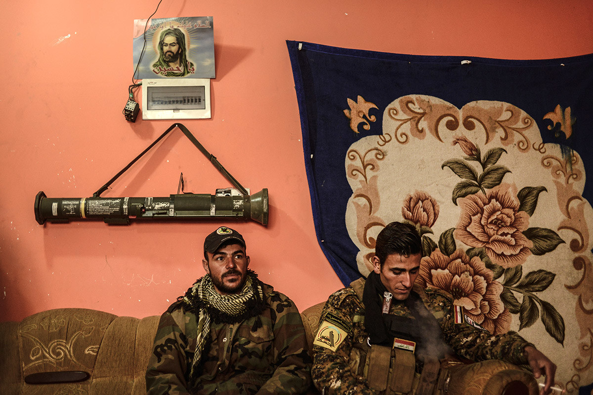 Shabak fighters Fouad (left), 27, and Ahmed, 25, of the Hashid Al-Shaabi's Babylon Brigade, rest after a patrol at their base on Jan. 28, in Bartella, Iraq. The Shabak are a persecuted minority group in Iraq who are targeted by Islamic State. Behind them hangs a picture of the Shia prophet, Imam Hussein, and a rocket-propelled grenade launcher.