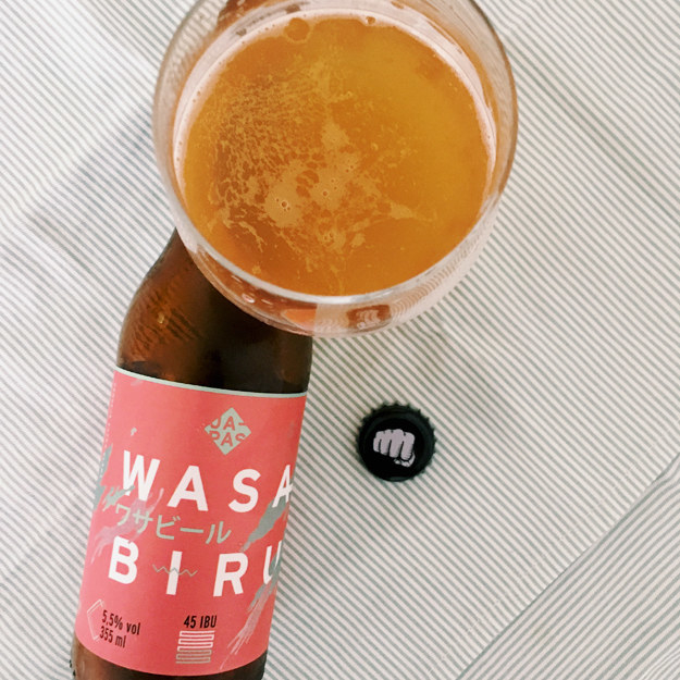 Eu adoro experimentar cervejas com ingredientes inusitados, e no mês passado, ganhei de presente de uma amiga esta cerveja com wasabi produzida aqui no Brasil.Ela é feita pela cervejaria JAPAS, comandada só por mulheres. A primeira leva da Wasabiru, uma American Pale Ale com 5,5% de teor alcóolico, foi produzida em 2014, mas eu ainda não havia experimentado e fiquei bem curiosa, já que wasabi não tem um sabor e nem um aroma que passa, digamos assim, despercebido. Logo ao abrir a garrafa já dá pra sentir um aroma forte, cítrico e refrescante que vem tanto do wasabi como do lúpulo japonês Sorachi Ace, usado na receita. Eu achei o sabor bem refrescante e equilibrado, os 45 IBU (unidade de amargor) podem parecer um pouquinho forte para alguns, mas nada demais comparados ao amargor de uma IPA. – Suria Rocha