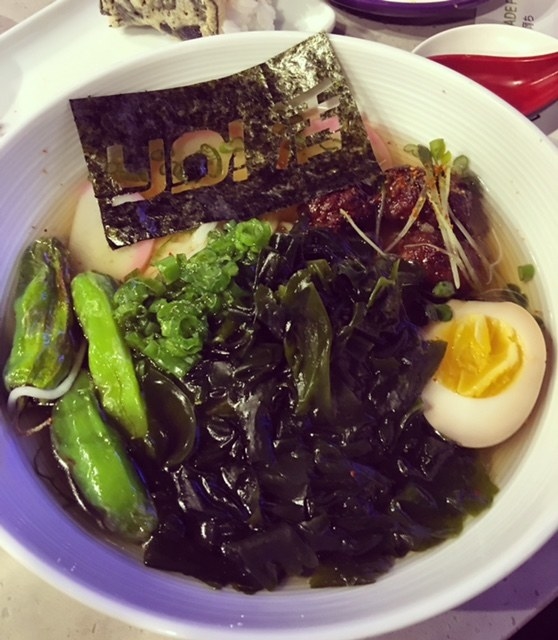A hearty bowl of ramen bowl filled with chicken tsukune, soft boiled egg, wakame, shishito peppers, kamaboko fishcake in a yuzu broth with nori garnish.