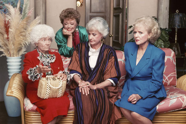 Three Golden Girls sitting on a couch and the fourth smiling and leaning on the back of the couch