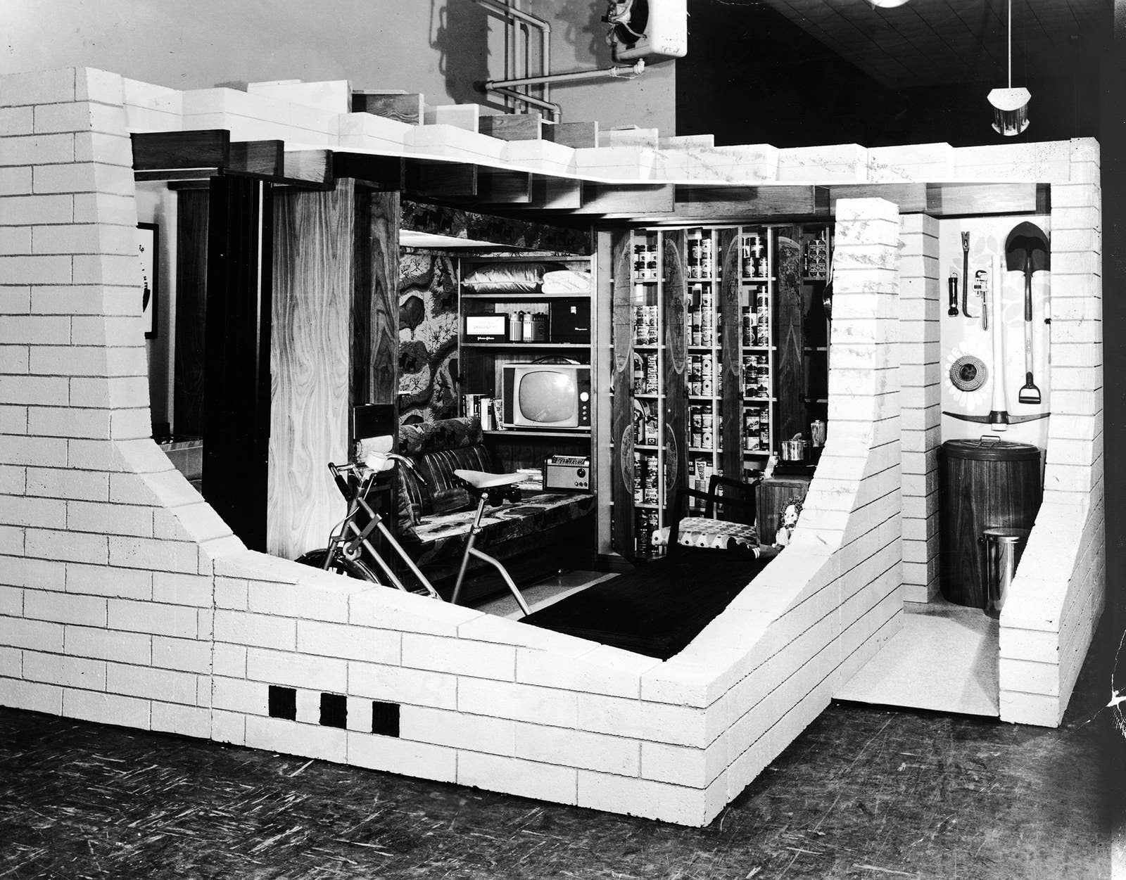 A cutaway display of a concrete masonry basement fallout shelter reveals an exercise bike, television, and library in the 1950s.