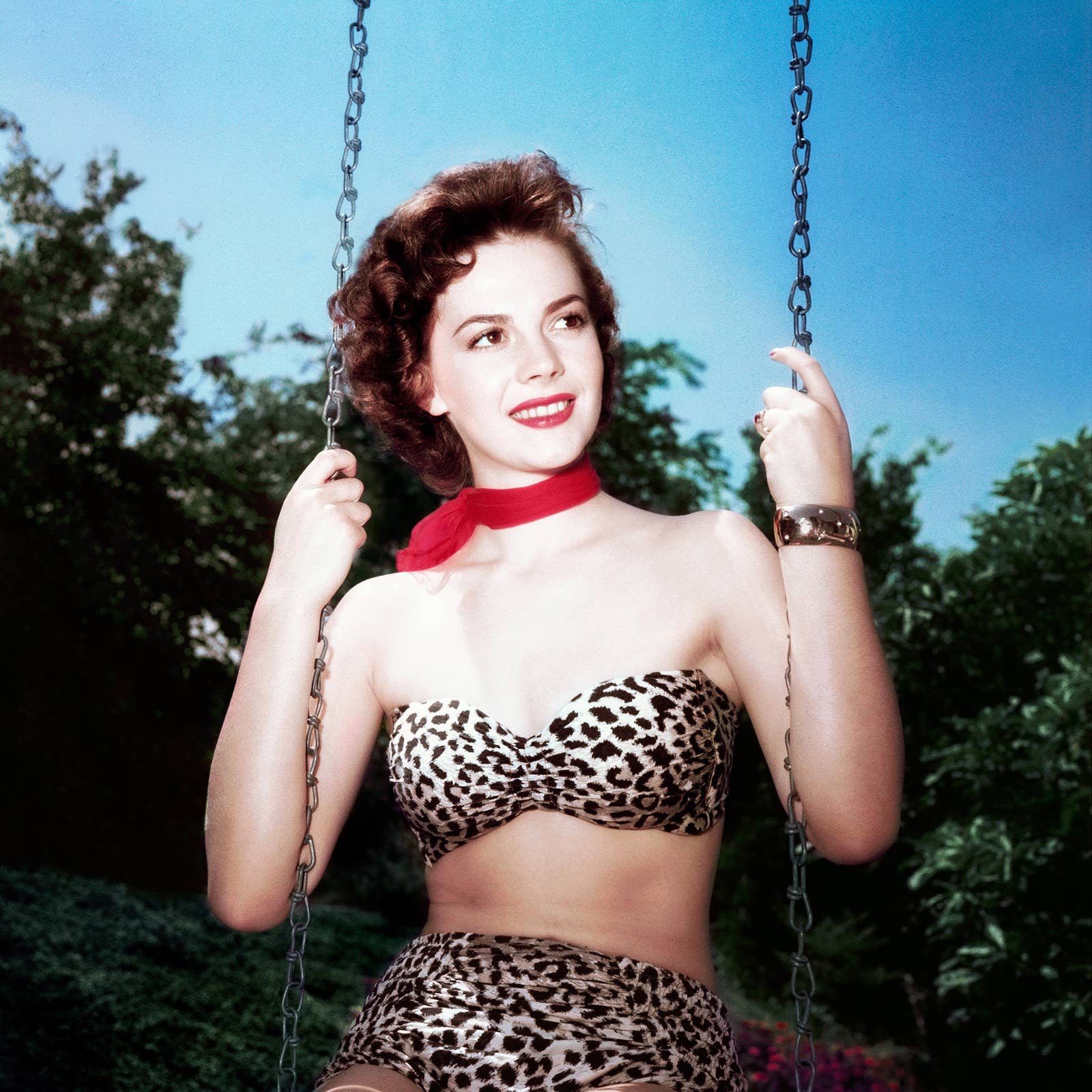 Actor Natalie Wood sits on a swing wearing a leopard-print bikini and a red...