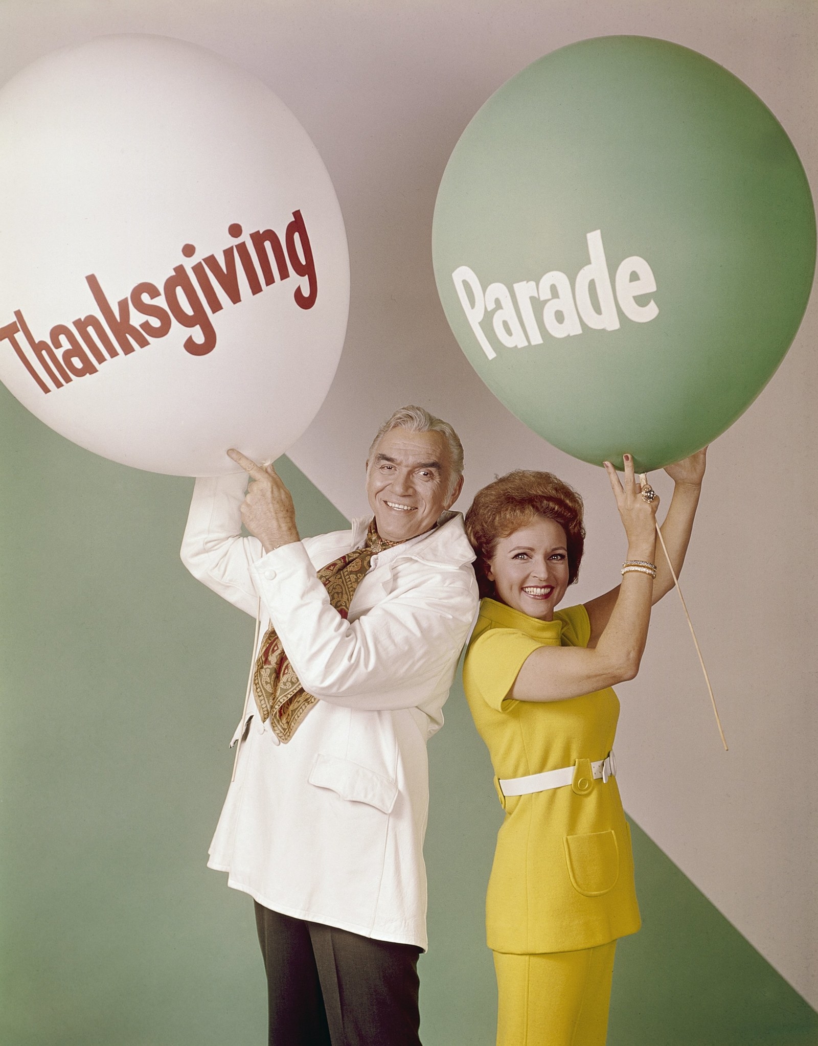 Smiling Lorne and Betty hold up balloons saying "Thanksgiving" and "Parade"