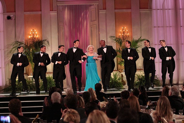 Betty onstage wearing a gown and standing in a line with many tuxedoed younger men