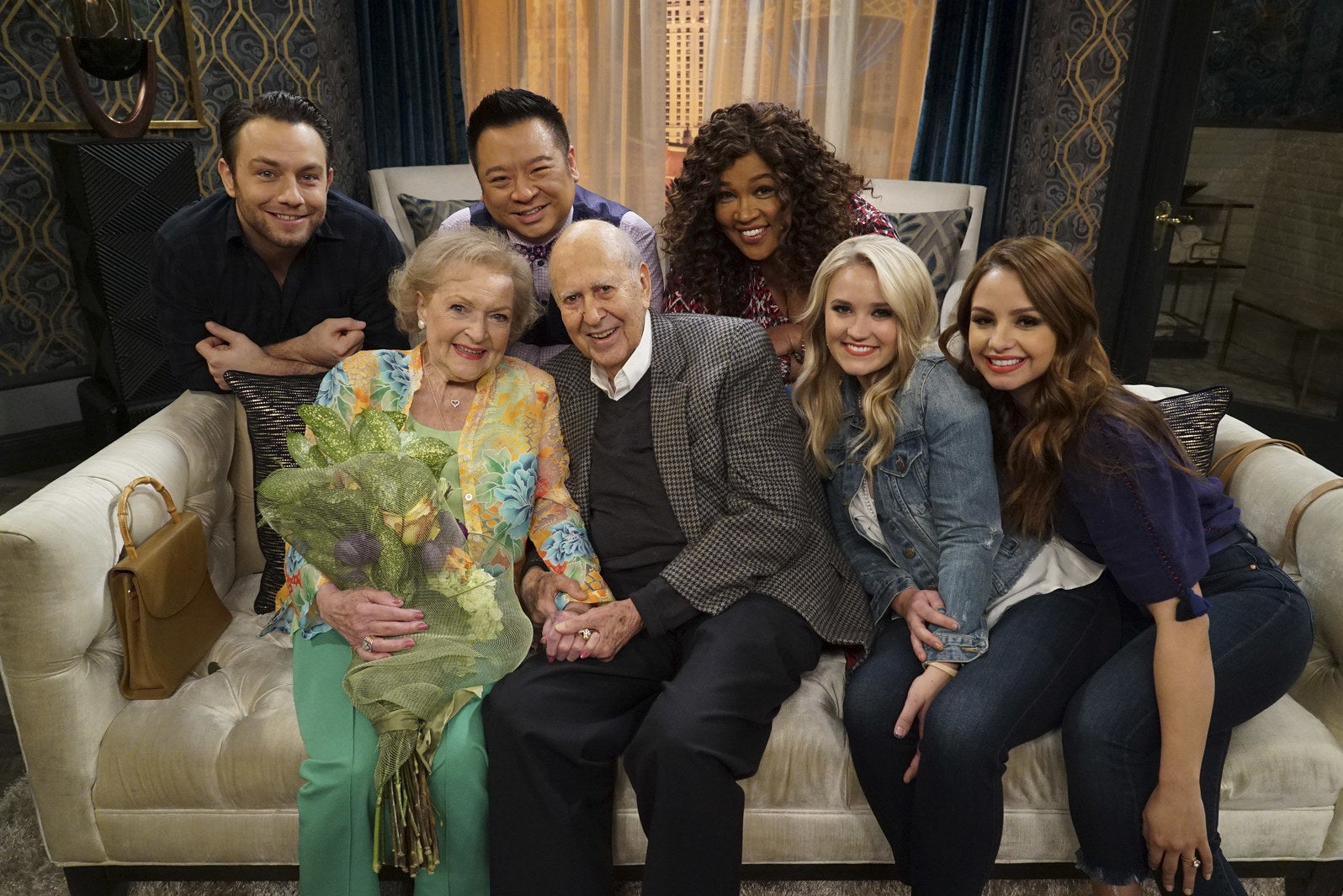 The cast, including smiling Betty holding a bouquet of flowers, at a couch