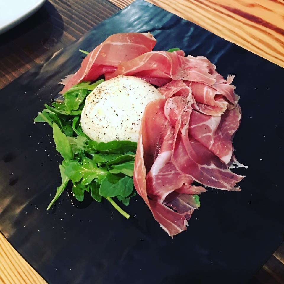 The buffalo mozzarella cheese served with arugula and prosciutto is the perfect antipasti to start your meal. The dish is garnished with a drizzle of olive oil and black pepper.