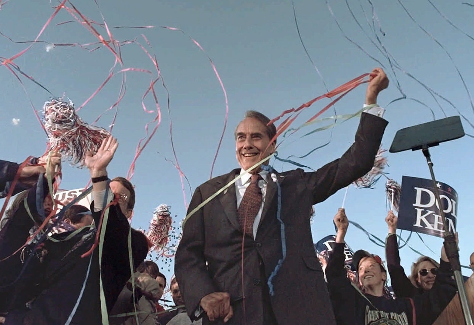 A smiling Bob Dole raises his left arm amid people waiving ribbons and banners