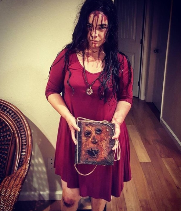 Someone in a red dress with blood on their face, holding a scary book