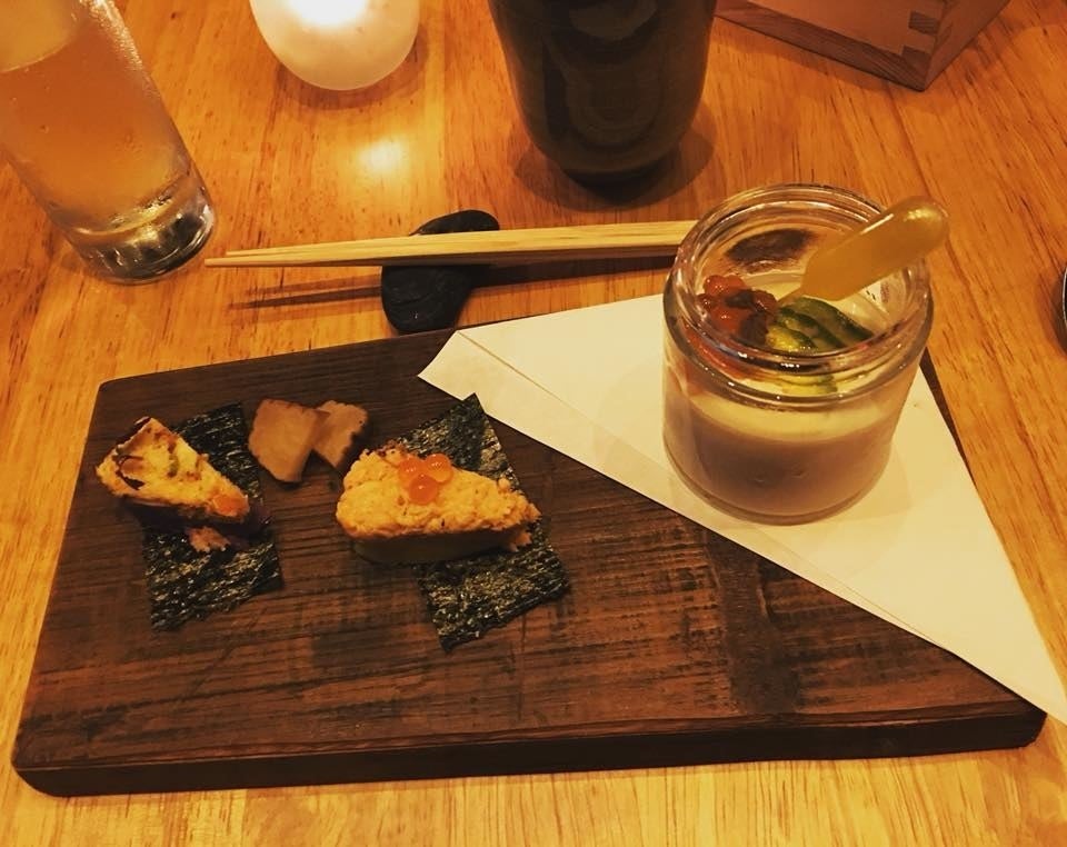 Served on a wooden platter are bite-sized Causa "Onigiri" - one is spicy crab meat on top of purple potato causa, while the other is spicy salmon covered in flavorful furikake with ikura on top of yellow potato causa. On the right is a bowl of pudding-like House Crafted Tofu with bonito flakes, Dashi Amarillo, ikura, oba, and avocado slices.
