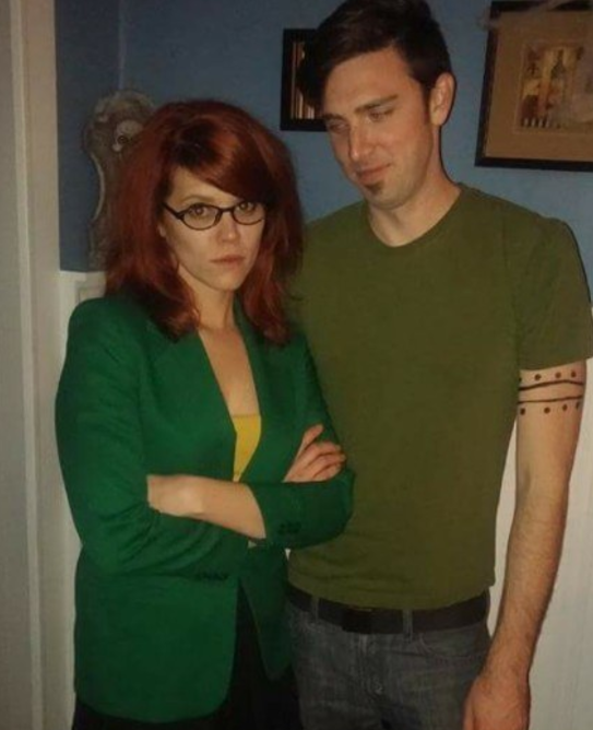 Woman in a suit jacket and long red hair and arms folded next to a man in a T-shirt