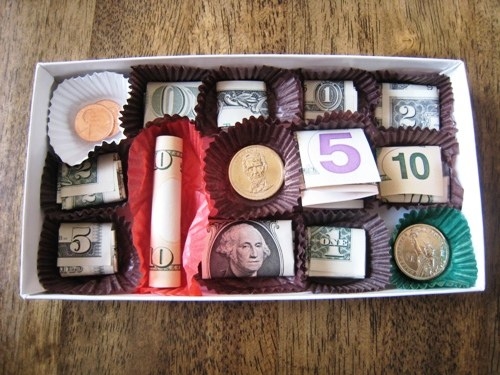 Open chocolate box with folded and rolled dollar bills inside accordion chocolate wrappers