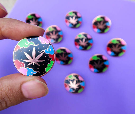 A colorful enamel pin featuring smoke clouds and a pot leaf.