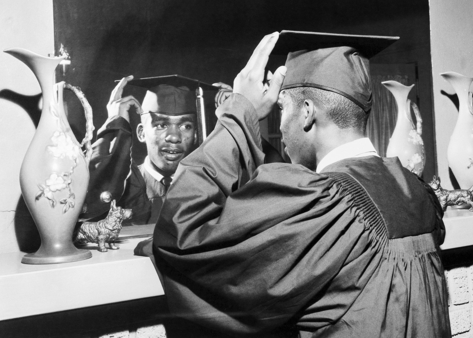 A Black boy wearing a cap and gown looking at himself in the mirror