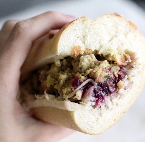 A hand holding a sub roll with roasted turkey, stuffing, cranberry sauce, and mayo