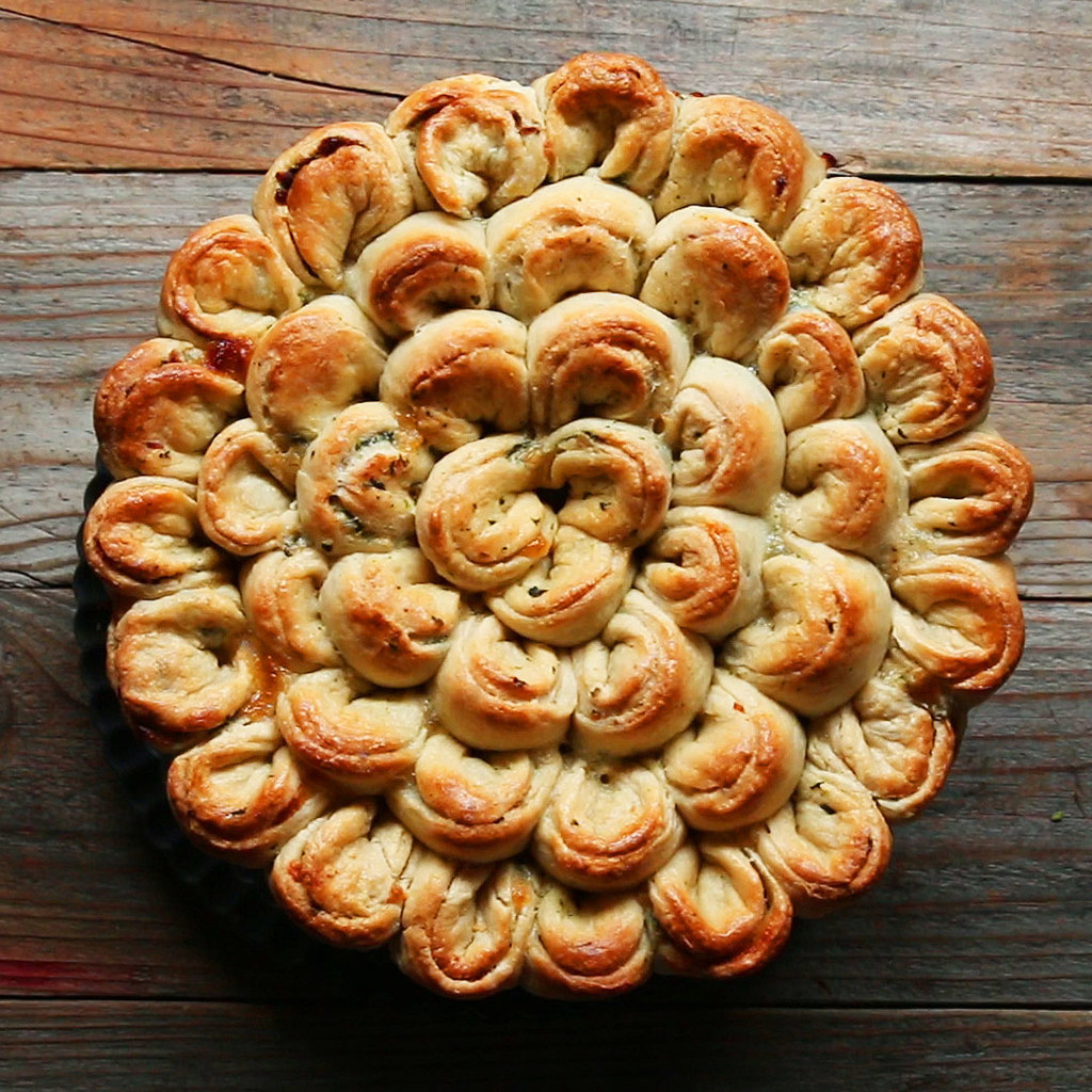 15 porçõesVocê vai precisar de: 230g de frango assado280g de cream cheese (queijo cremoso), amolecido20g (⅔ xícara) de espinafre, cozido e escorrido250g (2 xícaras) de queijo mussarela ralado1 colher de chá de alho em pó1 colher de chá de pimenta1 colher de chá de sal500g (4 xícaras) de farinha de trigo4 colheres de sopa de açúcar4 colheres de sopa de fermento em pó2 colheres de chá de sal12 colheres de sopa de manteiga sem sal, gelada480ml (2 xícaras) de leite integralMODO DE PREPARO1. Preaqueça o forno a 200°C.2. Peneire a farinha, o açúcar, o fermento em pó e o sal (tudo junto) em uma tigela grande. Corte a manteiga sem sal em pequenos pedaços e coloque-a na tigela (ou use um ralador de queijo para ralar a manteiga em pequenos pedaços). Use um garfo para incorporar a manteiga à mistura de farinha, até formar uma textura farelenta.3. Acrescente o leite e mexa a massa até que ela fique espessa e comece a se soltar das laterais da tigela. Transfira a massa para uma superfície plana, polvilhada com farinha. Sove a massa até ela ficar com cerca de 2 centímetros de espessura. Dobre-a no meio e sove-a novamente até chegar aos 2 centímetros de espessura. Cubra com uma toalha e deixe descansar por 30 minutos.4. Enquanto a massa descansa, misture o frango, o cream cheese, o espinafre, o queijo mussarela, o alho em pó, a pimenta e o sal em uma tigela grande.5. Polvilhe um rolo de massa com a farinha e abra a massa até ela ficar com ½ centímetro de espessura. Use a boca de um copo para fazer 45 círculos na massa.6. Remova a pasta de frango e espinafre da geladeira e coloque 1 colher da pasta em cada círculo de massa. Dobre os círculos no meio e pressione as laterais para selar a pasta dentro da massa. Dobre e encoste o lado esquerdo no lado direito de cada semicírculo, como em um biscoito da sorte.7. Coloque a massa recheada em uma forma com fundo removível (antiaderente ou já untada) de 28 centímetros. Faça um círculo externo e trabalhe de fora para dentro, criando 4 círculos no total.8. Leve ao forno por 25 minutos ou até dourar.9. Sirva e bom apetite!10. Inspirado em: https://www.liveinternet.ru/users/3560462/post337895407/