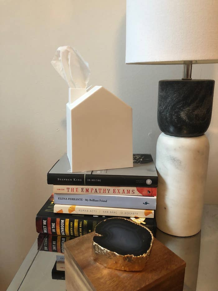 night stand table with a stack of books, lamp, then a square shape house-like white tissue box cover