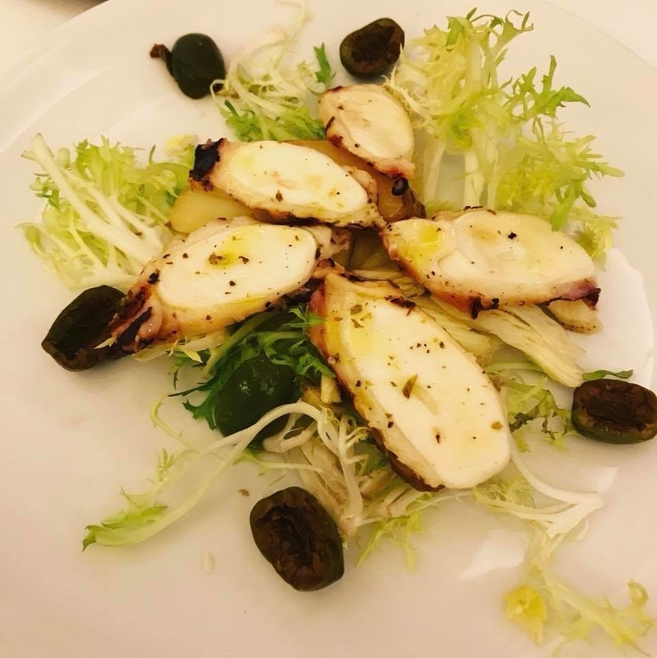 Perfectly prepared grilled octopus is served with frisée, celery hearts, oven-baked fingerling potatoes, and Castelvetrano olives. The dish is topped with a light drizzle of extra virgin olive oil and freshly ground pepper.