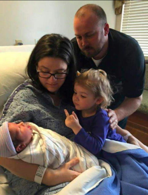 A mother holds a newborn in her arms while her toddler sitting in her lap gives the baby the middle finger, and the father looks on