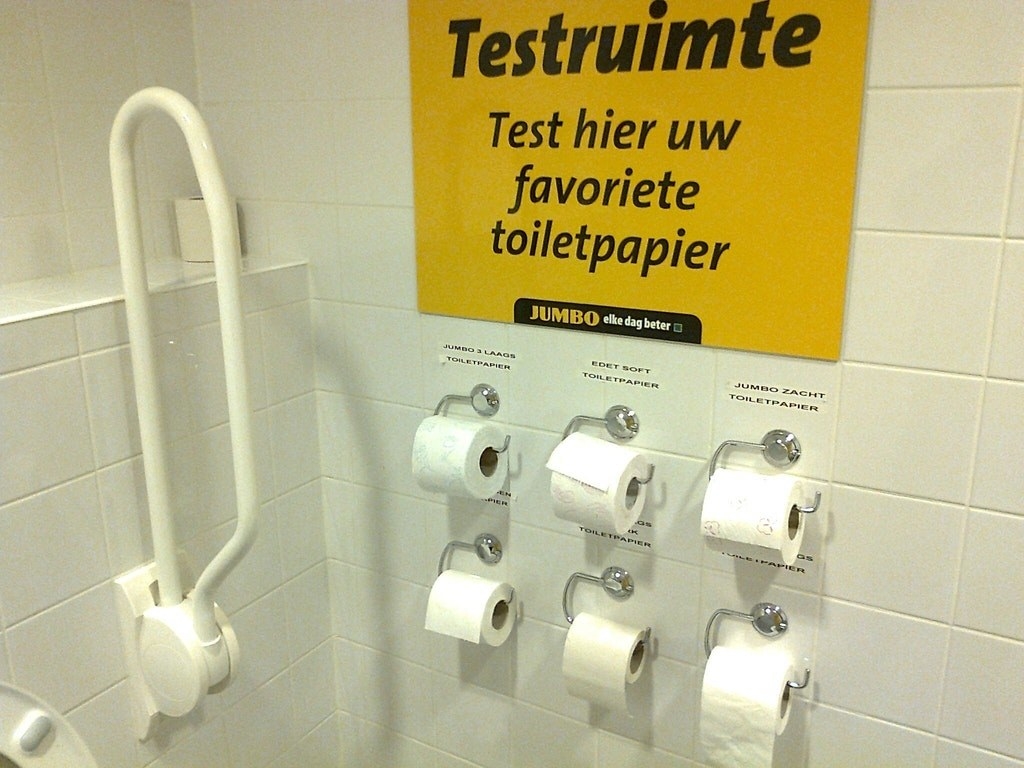 A wall of a public bathroom with six toilet paper rolls mounted to the wall, each labeled with a different brand so customers can know what they&#x27;re trying.