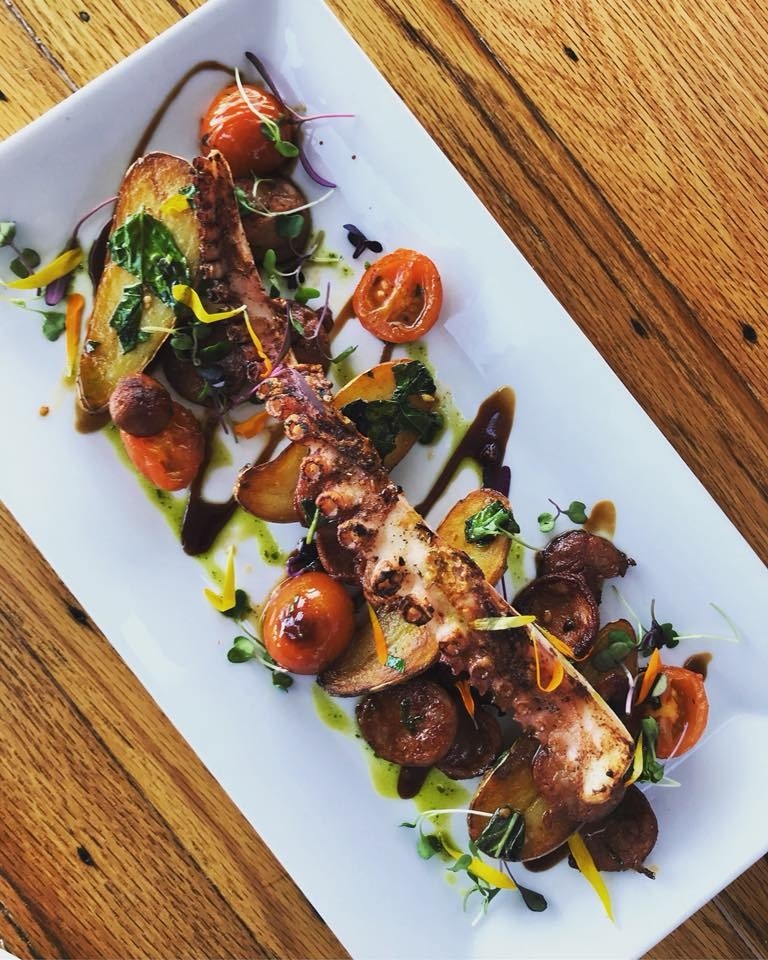 A piece of plump, juicy octopus is complemented by slices of chorizo, baby potatoes, and garlic fresh tomato. The dish is drizzled with pesto olive oil and vinaigrette.