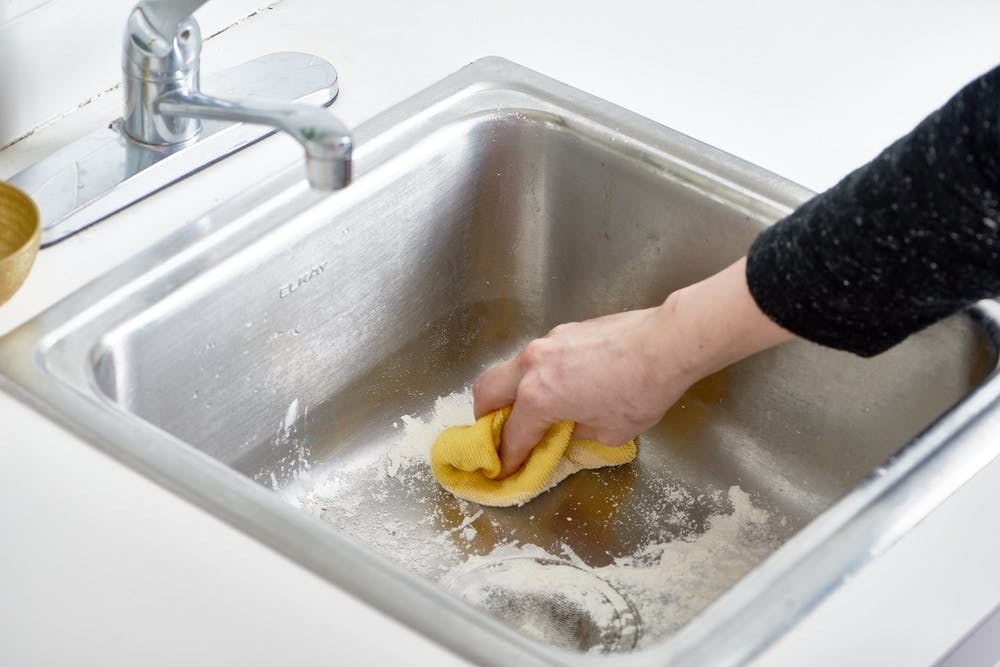 A person using flour and a microfiber cloth to make their stainless steel sink look shiny and clean