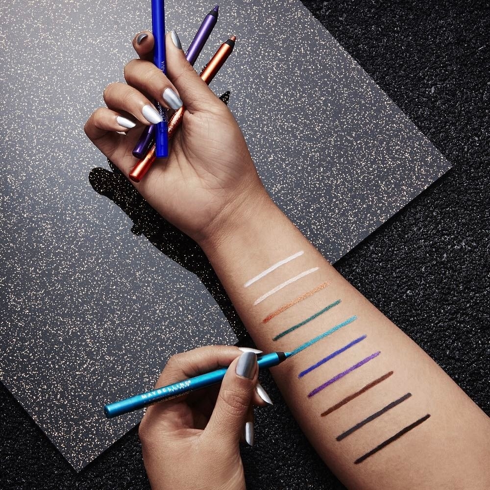 An arm holding an assortment of the Maybelline eyeliner, plus swatches of 10 different shades, including metallic colors and neutrals like black and brown
