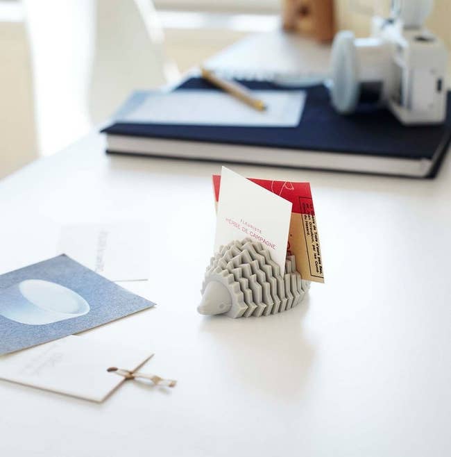 Hedgehog-shaped card holder on a desk, displaying cards, with books and writing tools in the background. Suitable for office or home decor