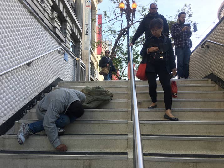 A man lies on the steps of the Powell Street BART station as evening commuters walk by.