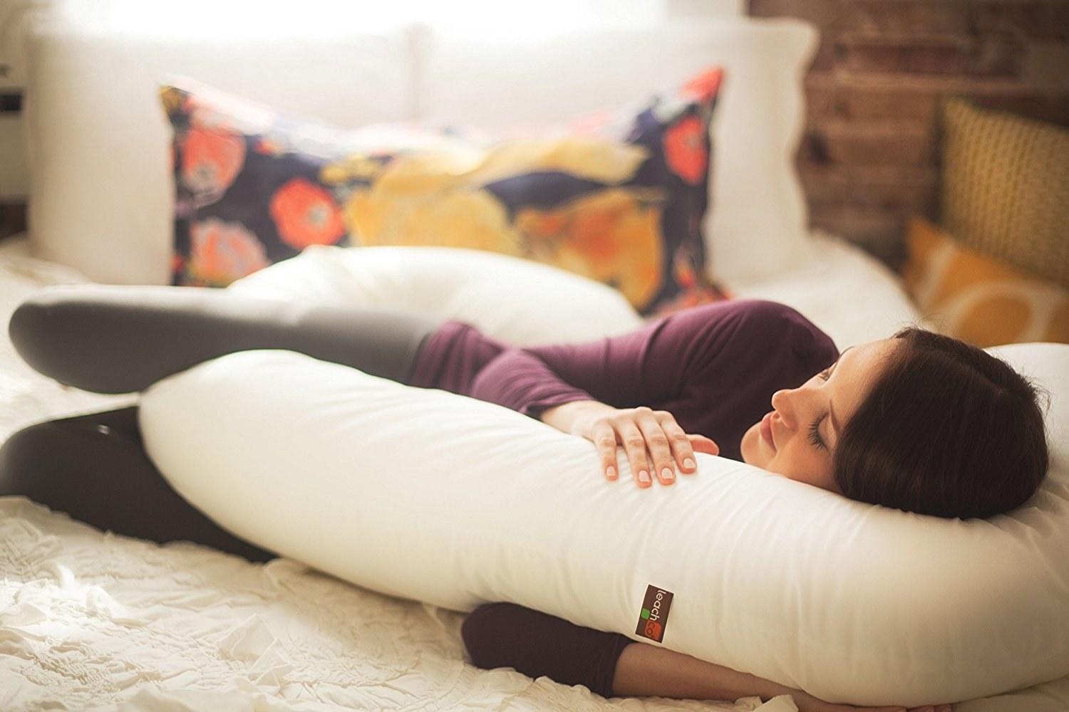 A model spooning the curved body pillow. Their back is supported by one end while their arms are around the other end and they have the curved bottom between their legs, elevating their knee. 