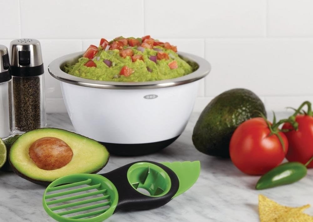 The green tool with a pitter, bladeless knife, and sliver next to a bowl of guacamole