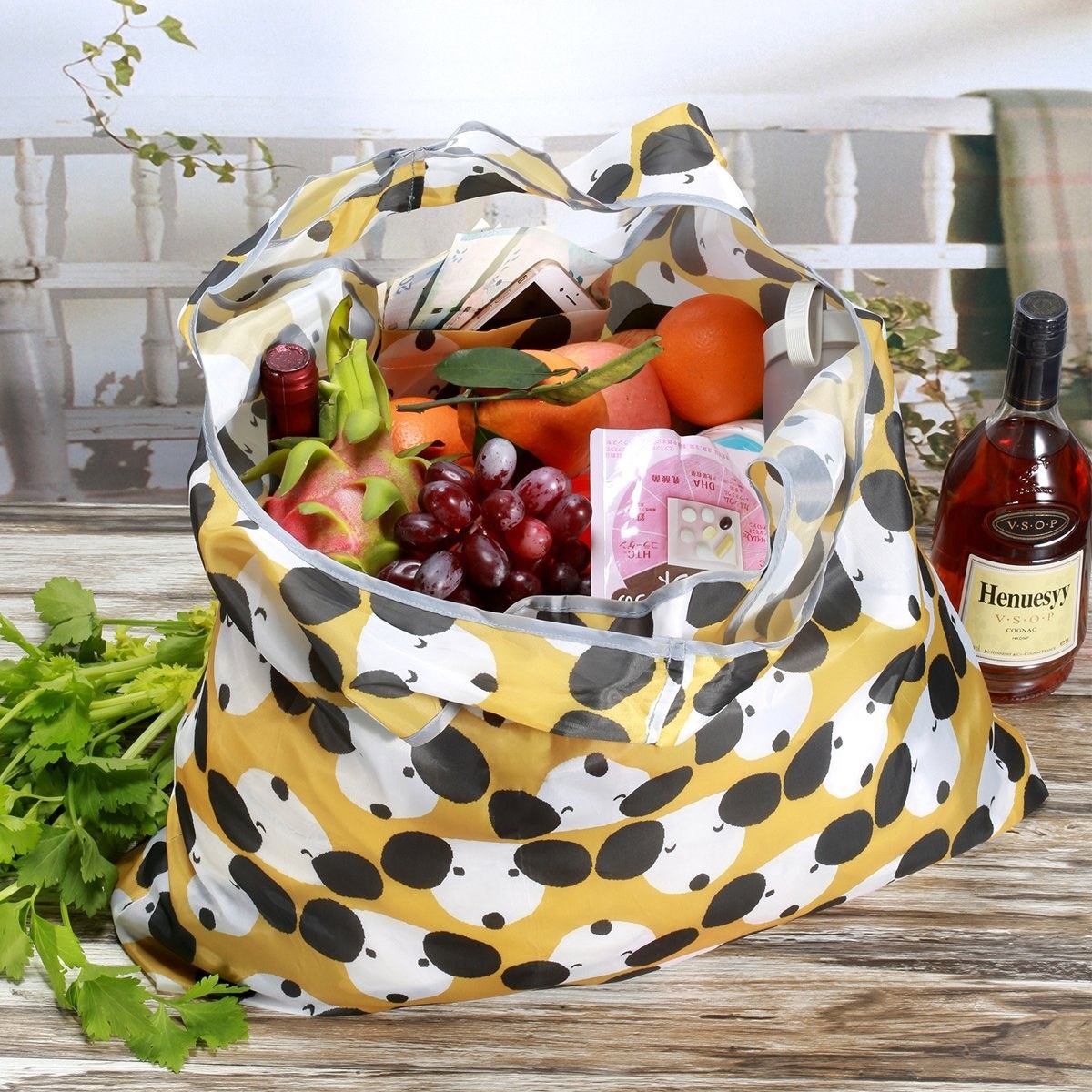  A yellow reusable bag with a handle with a dog print and holding groceries, including a wine bottle, grapes, oranges, and other items